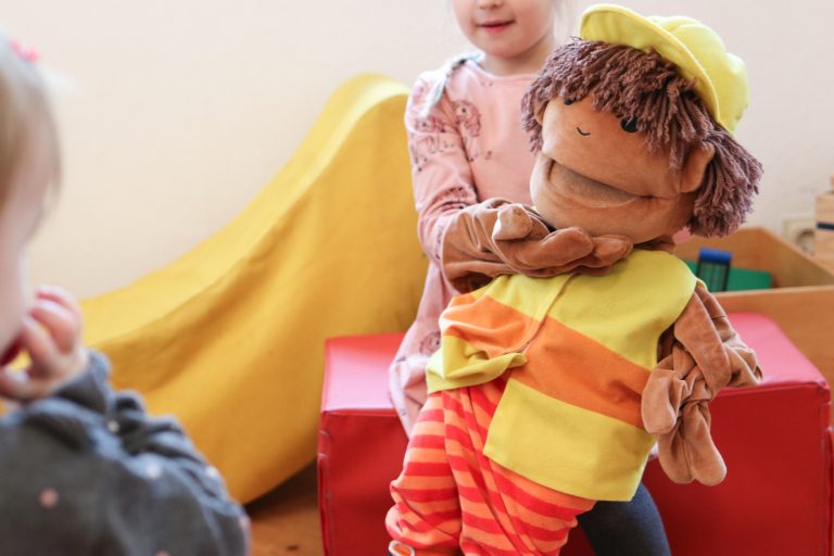 Ein junges Mädchen mit rosa Kleid spielt mit einer Handpuppe und ein jüngeres Mädchen schaut ihr dabei zu.