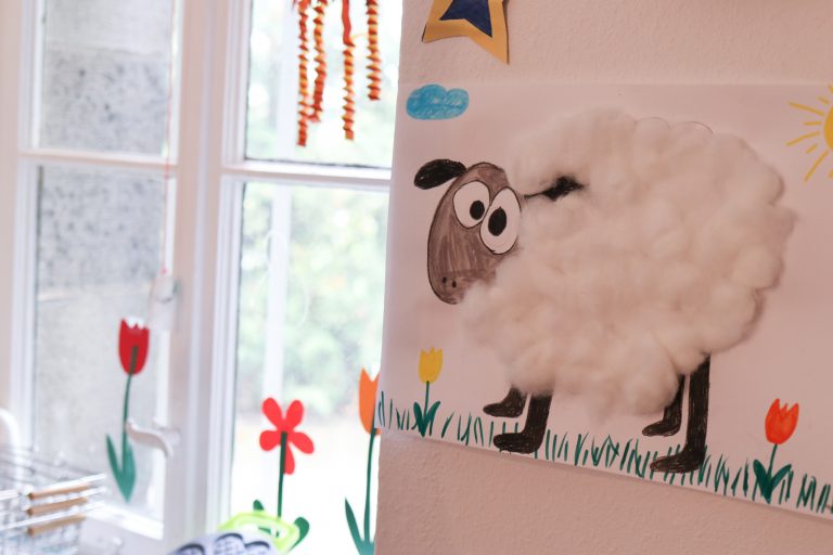 An einer weißen Wand neben einem Fenster hängt ein gemaltes Bild eines Schafes mit aufgeklebter Watte als Schafspelz.