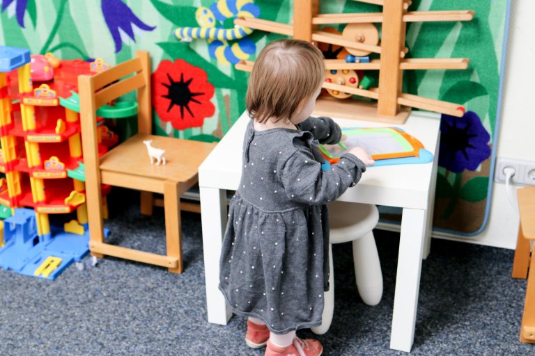 Ein kleines Mädchen in grauem Kleid spielt in der Spielecke an einem weißen Tisch